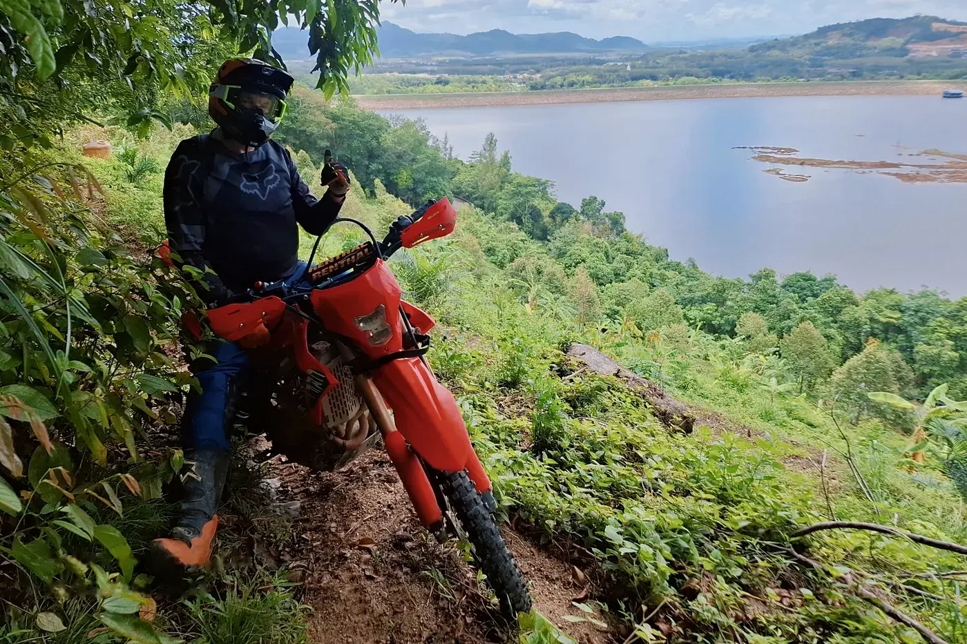 A rider gives a thumbs up from a red Honda CRF300L on a narrow hillside trail above a reservoir in Phuket
