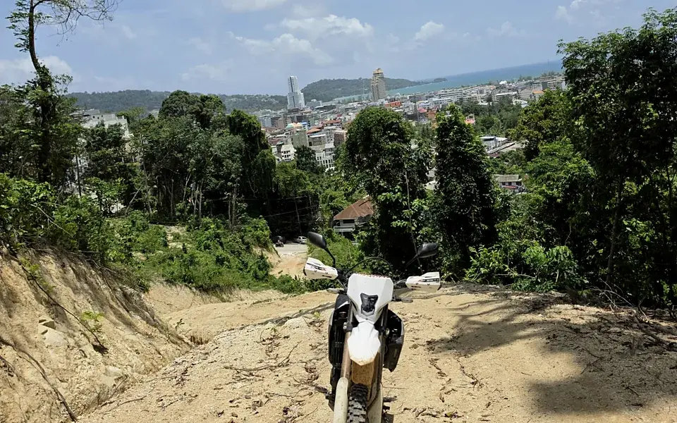 A dirt bike stopped at the top of a sandy trail, with a town and the sea below