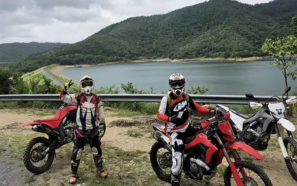 Two riders in full gear stopped with their bikes above a lake, one giving a thumbs up