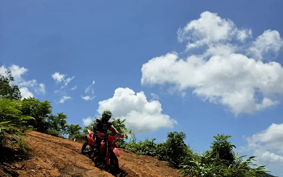 A rider on a red dirt bike on a clay slope, under a blue sky with big white clouds