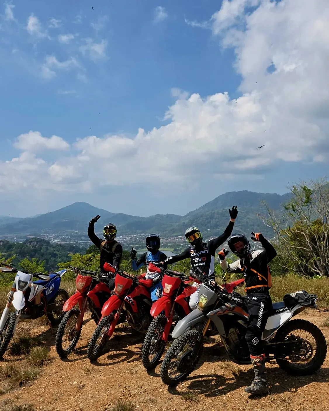Four riders in full gear wave from a hilltop above Phuket, their dirt bikes lined up in front of them