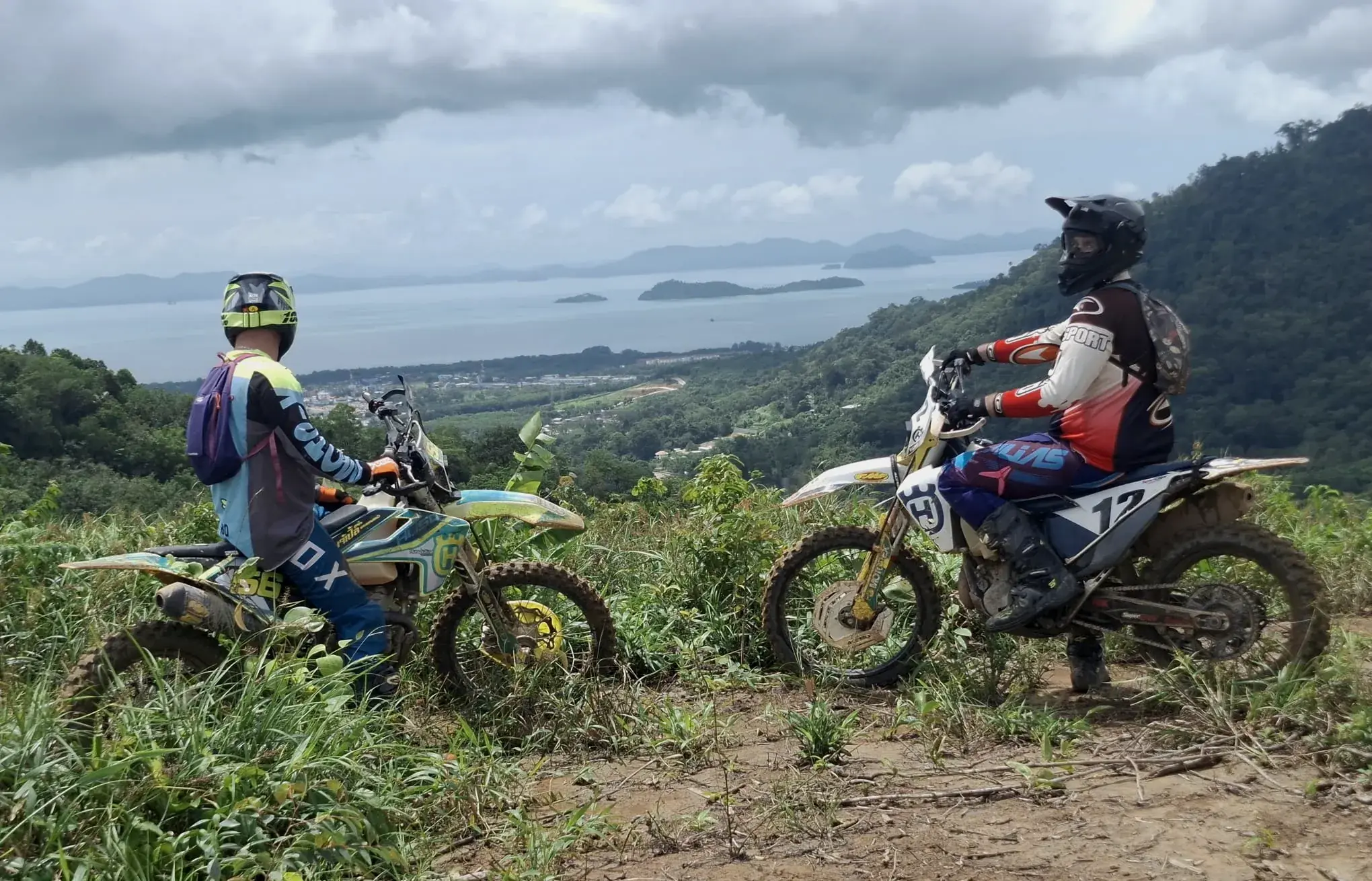 Two riders stop their dirt bikes on a hilltop trail above the bay off Phuket