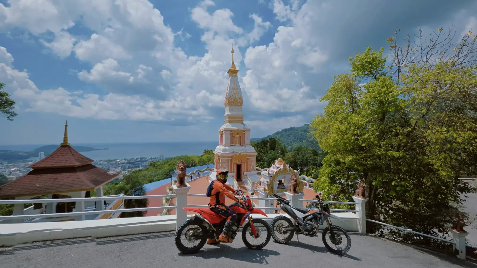 A rider in orange gear on a red dirt bike beside a hilltop temple, with green hills and the sea behind, in Phuket