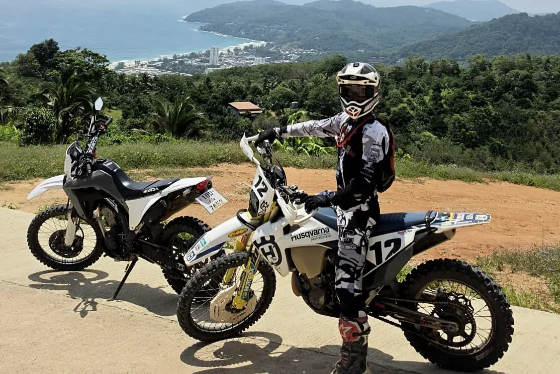 A rider in full gear beside a white Husqvarna enduro bike, number 12, at a hilltop viewpoint above a Phuket beach, a second bike parked behind