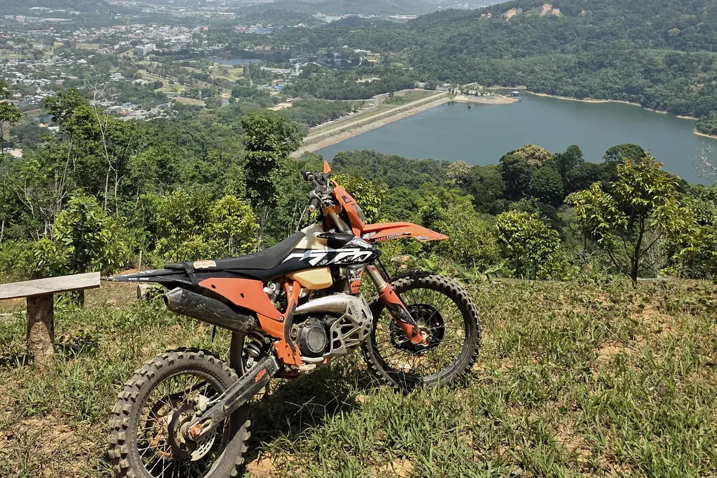An orange KTM enduro bike parked on a grassy hilltop above a reservoir in Phuket