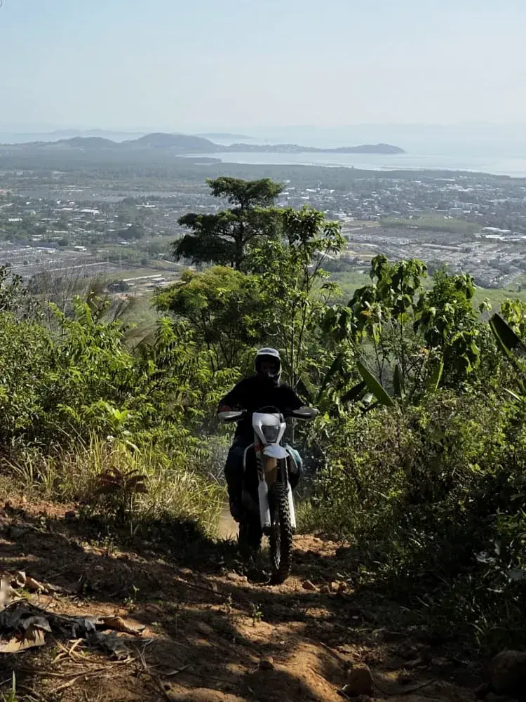 A rider follows a narrow ridge trail in Phuket, with the town and the sea far below