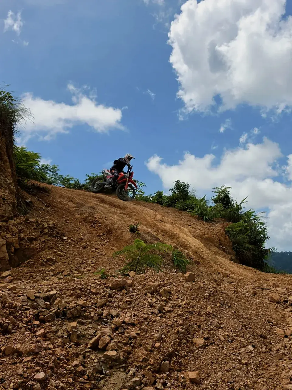 A rider tops a loose, rocky dirt climb under a blue sky in Phuket